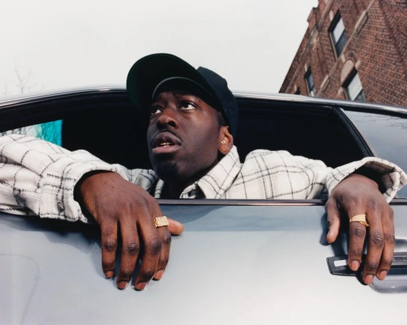 Portrait of a dark-skinned man in car, wearing baseball cap and gold rings, gazing upwards on an overcast day in an urban setting.