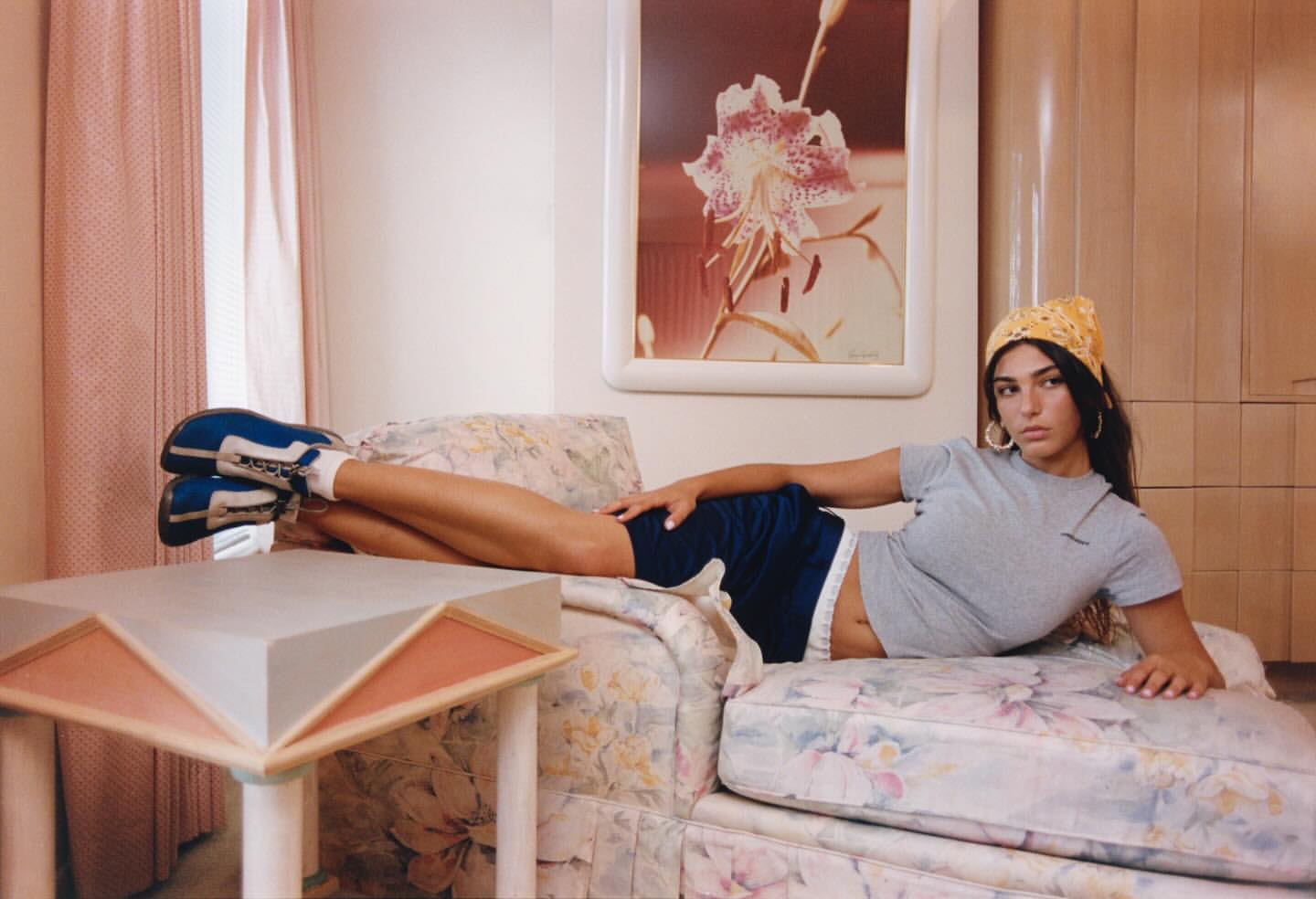 A woman lounges on a floral couch in a casual outfit, complemented by vintage athletic shoes and a yellow bandana in an indoor setting.