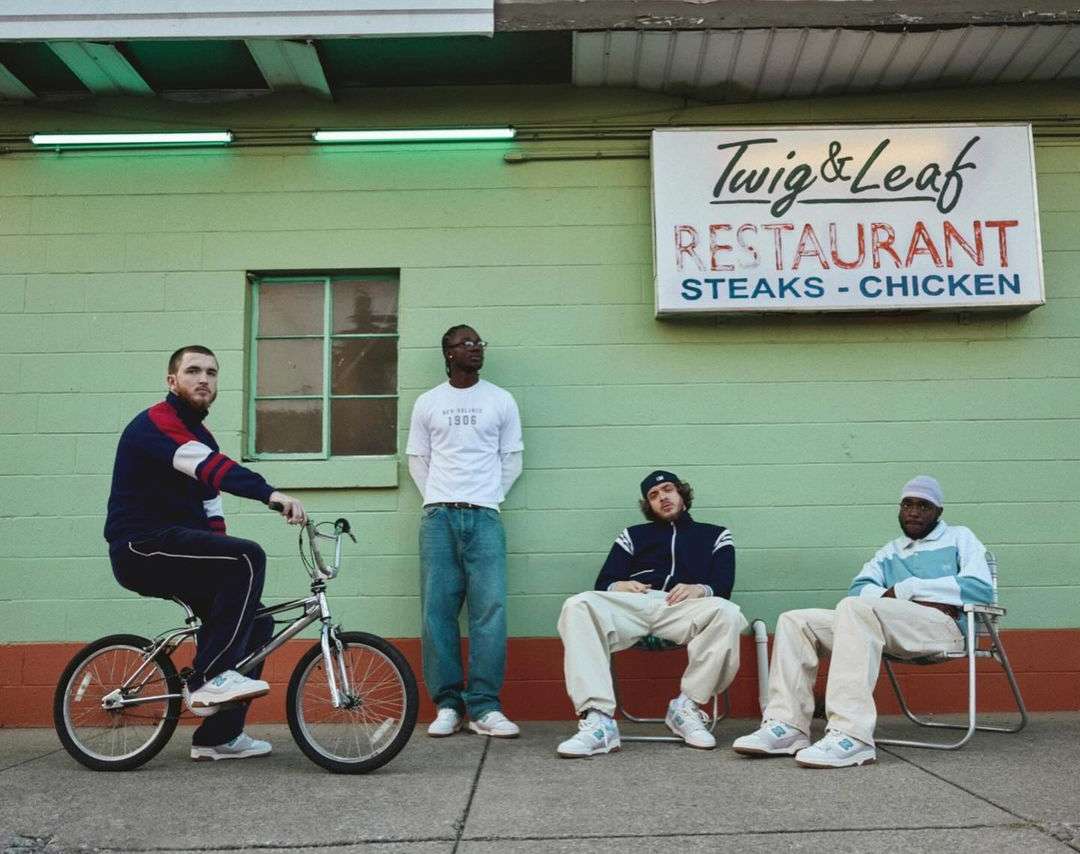 Four men pose outside a green Twig & Leaf restaurant, one riding a bicycle, all wearing casual clothing and sneakers.