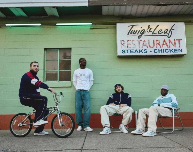 Four men pose outside a green Twig & Leaf restaurant, one riding a bicycle, all wearing casual clothing and sneakers.