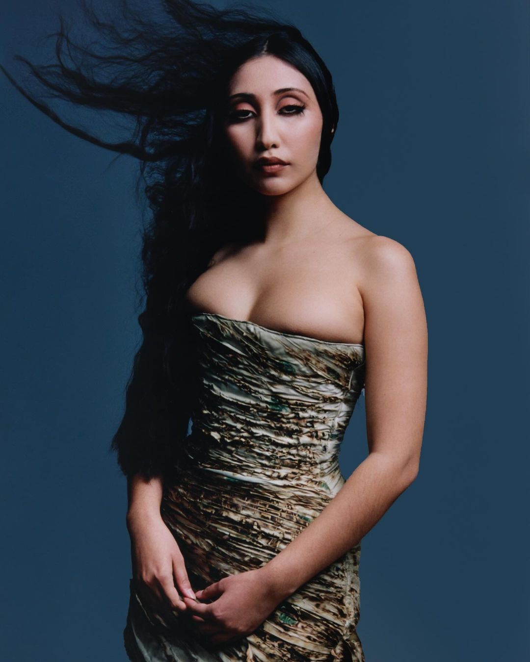 Portrait of a woman with long dark hair blowing in the wind wearing a strapless textured dress against a dark blue background.