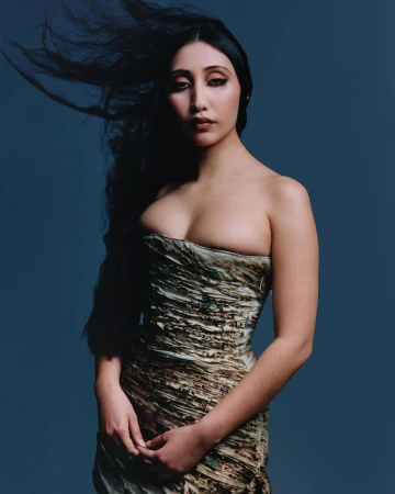 Portrait of a woman with long dark hair blowing in the wind wearing a strapless textured dress against a dark blue background.