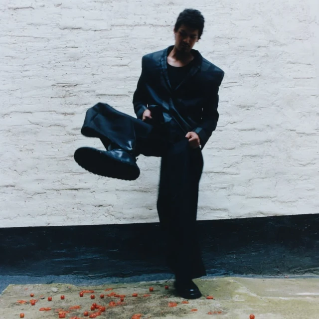 Fashionable person in black suit poses outdoors, kicking one leg up near a brick wall and scattered red candies on the ground.
