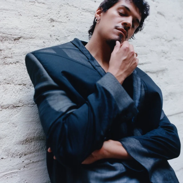 Adult male with a mustache in a dark blue textured jacket leaning against a white wall, close-up shot. Fashion photography.