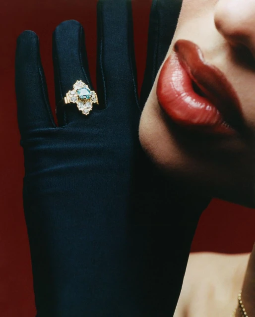 Close-up studio shot of a model wearing a black glove with a gold ring, set against a red backdrop, highlighting beauty and fashion.