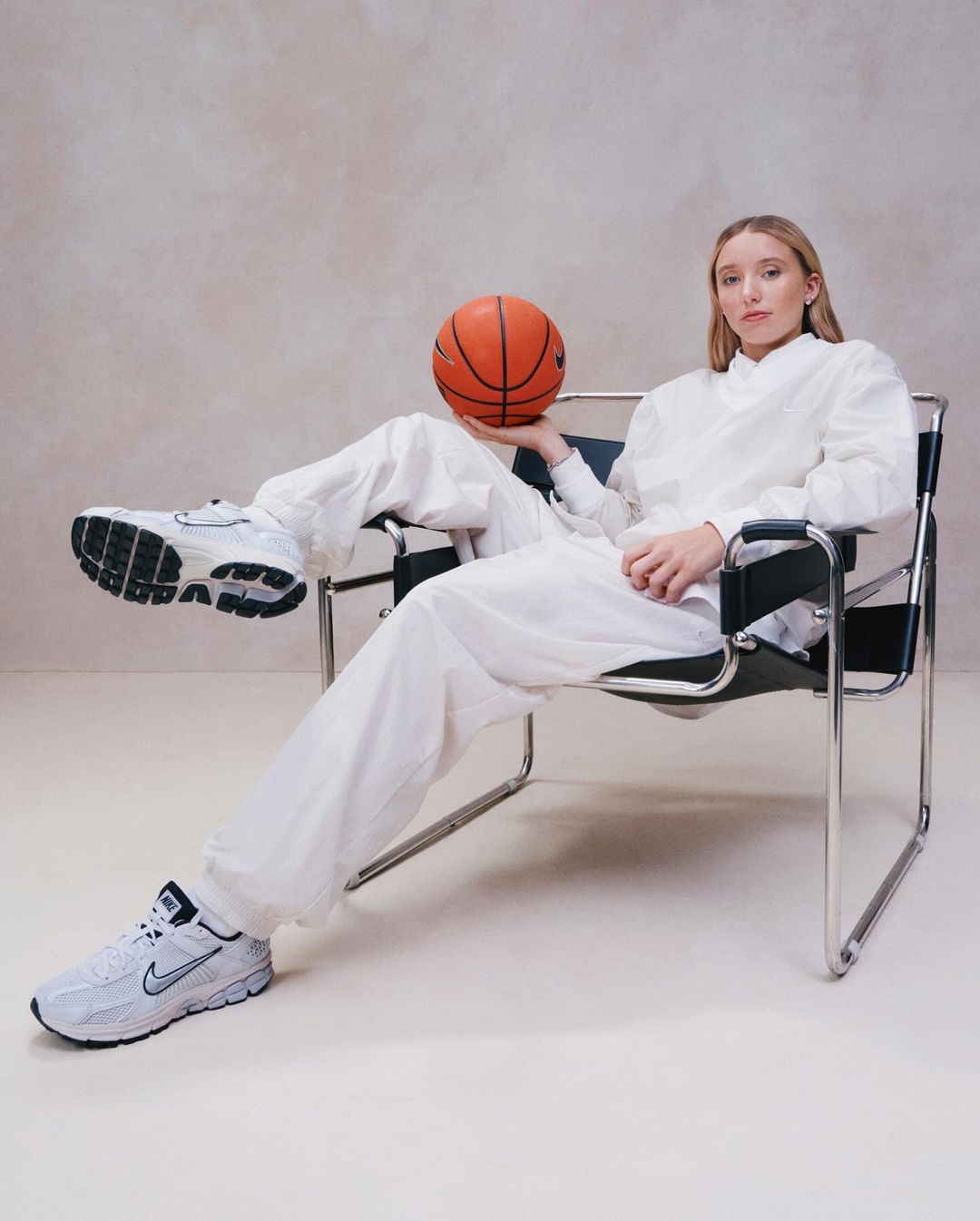 Young woman in a white outfit sitting in a chair, holding a basketball, with white Nike sneakers, in a neutral indoor setting.