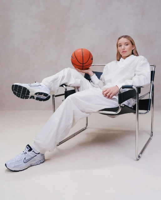 Young woman in a white outfit sitting in a chair, holding a basketball, with white Nike sneakers, in a neutral indoor setting.