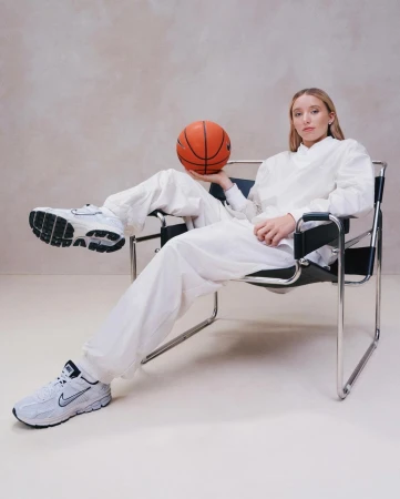 Young woman in a white outfit sitting in a chair, holding a basketball, with white Nike sneakers, in a neutral indoor setting.