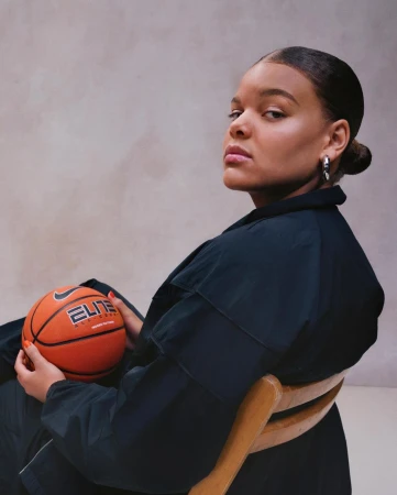 Woman in dark clothing holding a basketball while seated on a wooden chair against a neutral studio backdrop