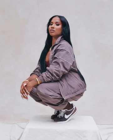 Woman with long dark hair wearing a mauve tracksuit and sneakers, squatting on a white block against a neutral backdrop.