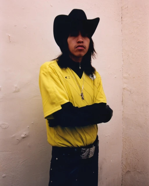 A person wearing a black cowboy hat, yellow t-shirt, and black undershirt poses in front of a wall with arms crossed looking at the camera.