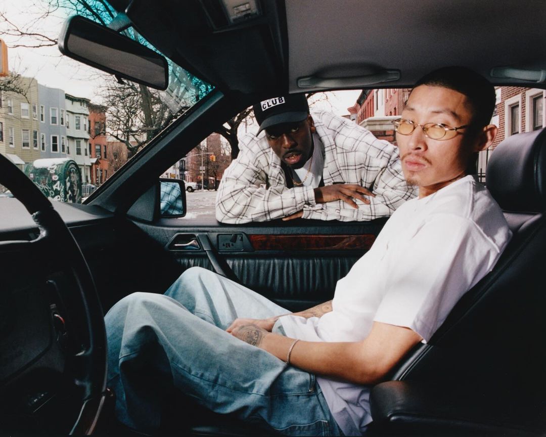 Two men posing inside and beside a vintage car in an urban street setting, photographed by Ricky Alvarez.
