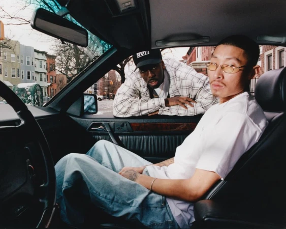 Two men posing inside and beside a vintage car in an urban street setting, photographed by Ricky Alvarez.