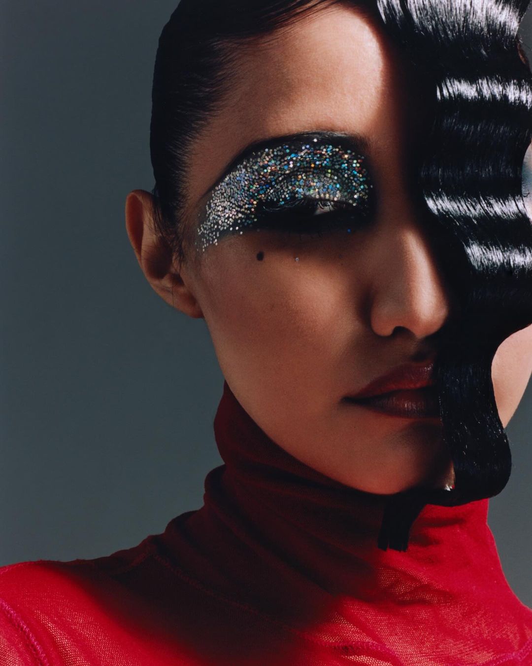 Close-up portrait of an adult woman with dark hair and red turtleneck. She has glittering makeup on her eyelid, shot in studio lighting.