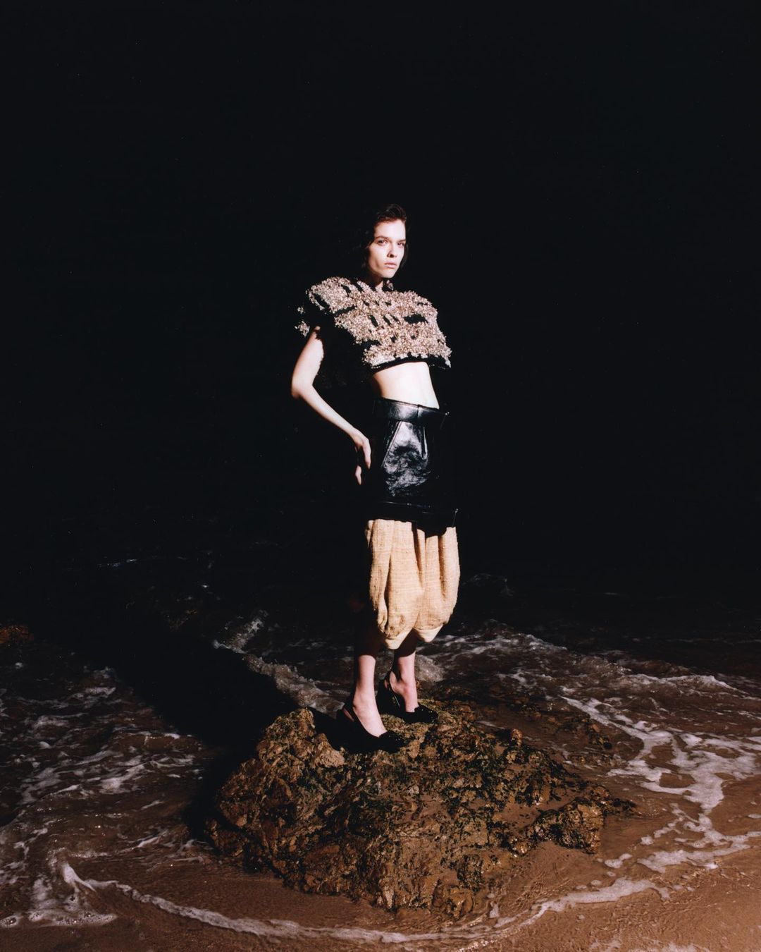 A woman in modern fashion stands on a rock surrounded by ocean water against a dark background. Her pose is confident and chic.