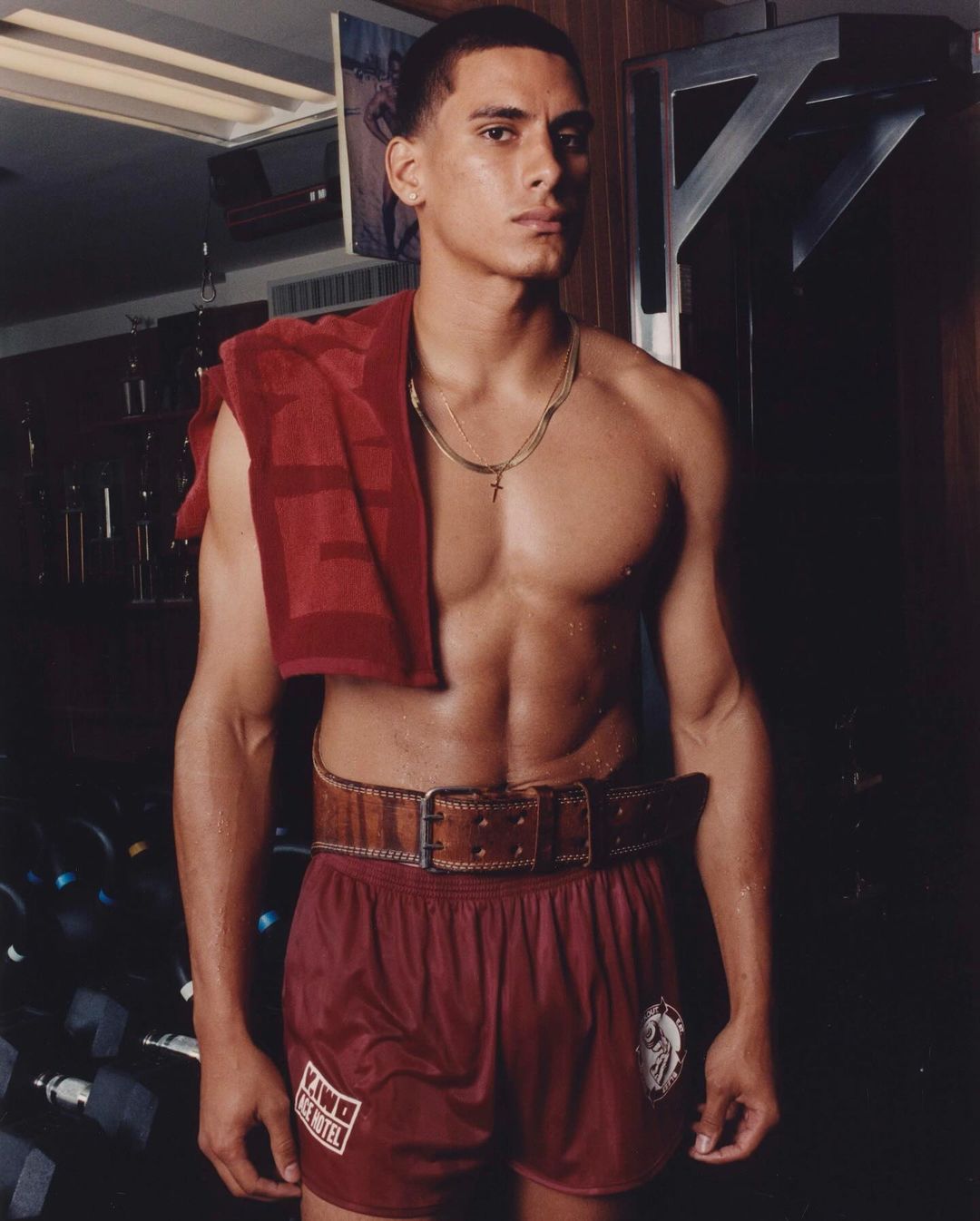 Shirtless man stands in a gym with a towel slung around his neck wearing red shorts with a wide belt with weights in the background.