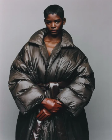 Model with short dark hair wearing a glossy quilted puffer coat and brown leather gloves against a plain grey background