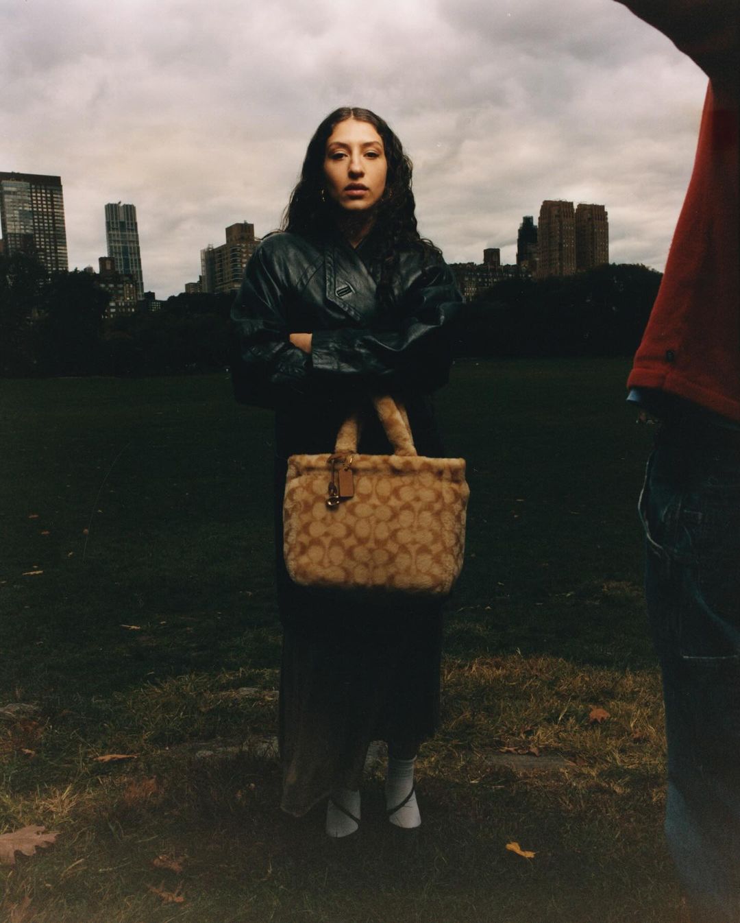 A young woman in a coat and handbag stands in a park with city buildings in the background, on a cloudy day, facing the camera.