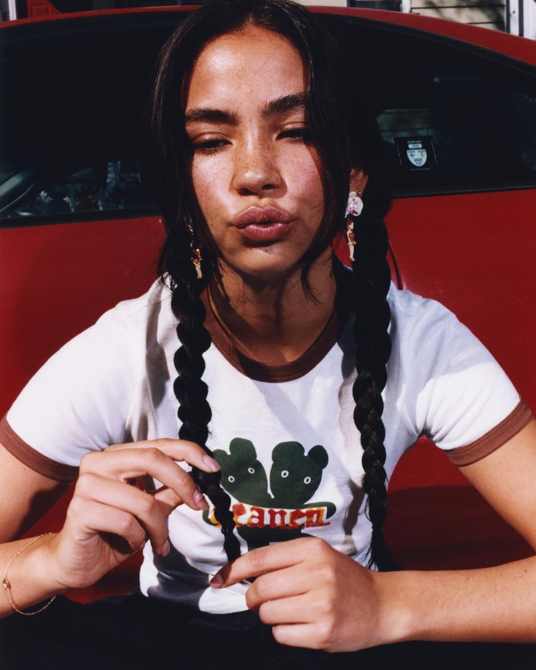 Young woman with dark hair in braids puckers lips while sitting in front of a red car, wearing a white shirt with graphic art.