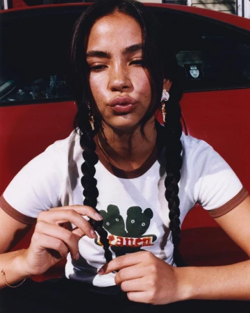 Young woman with dark hair in braids puckers lips while sitting in front of a red car, wearing a white shirt with graphic art.