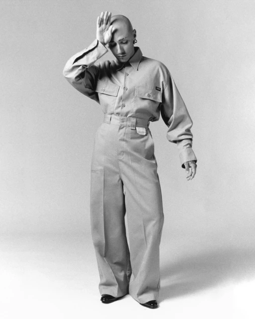 Full-length portrait of a bald adult in a tan collared shirt and wide-leg pants, hand on head, on a neutral backdrop.