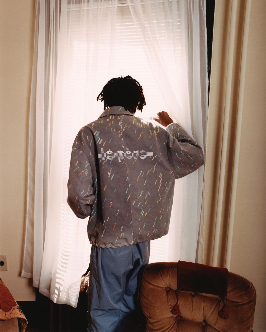 Adult standing at a window with sheer white curtains, wearing a patterned jacket and blue pants in an indoor setting with tan walls.