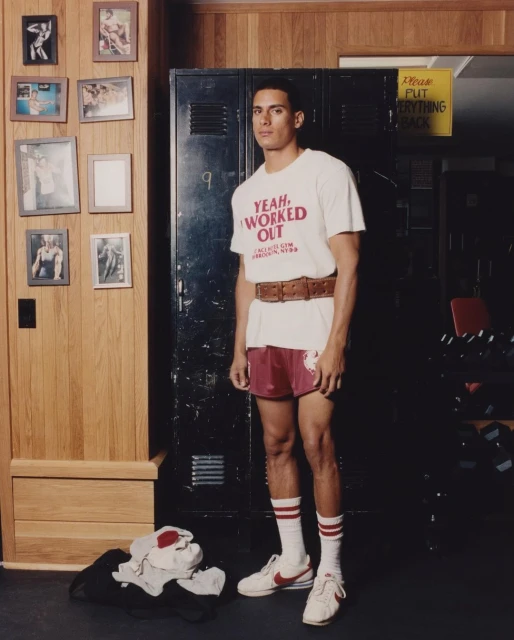Portrait of an adult male in athletic attire in a locker room with photos, wearing a t-shirt with a slogan, shorts, sneakers, and a belt.