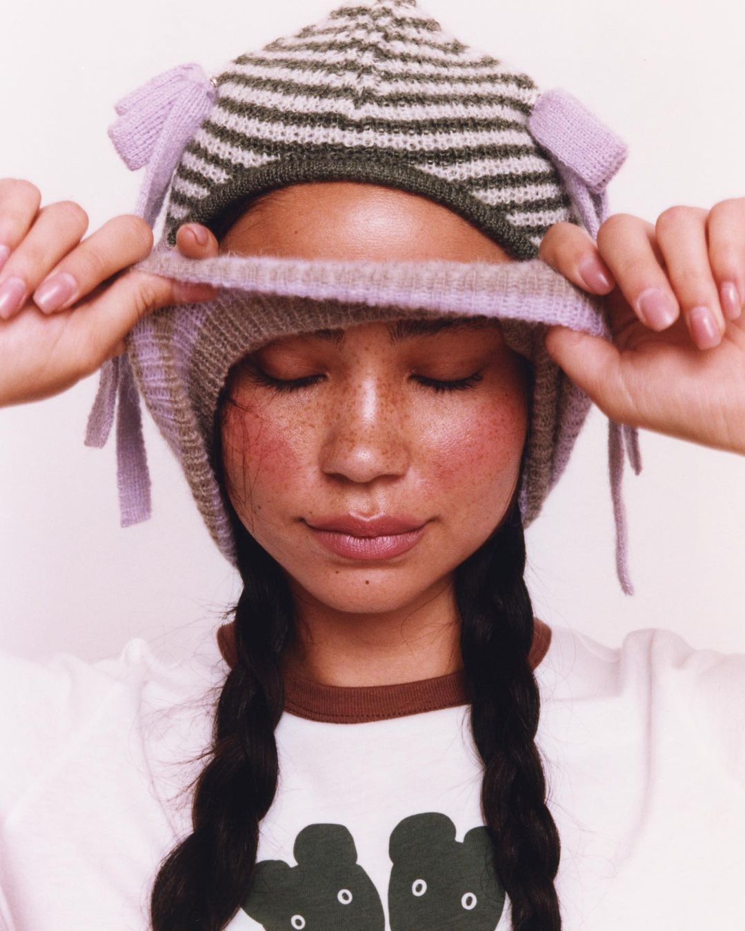 Close-up of a woman wearing a striped knit hat with tie details, hands adjusting the hat, closed eyes, and a white T-shirt.