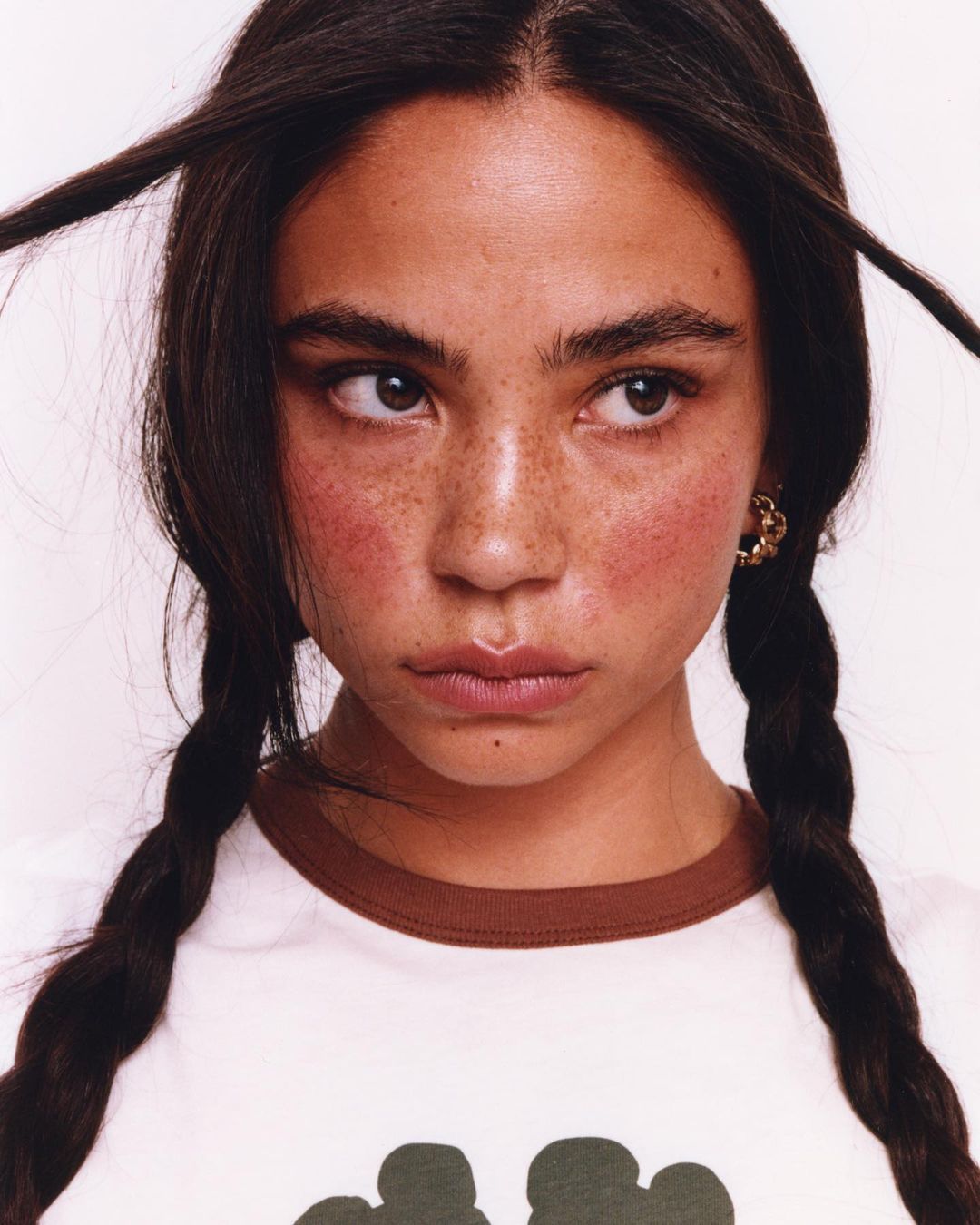 Close-up of a young woman with braided brown hair, fair skin, and freckles, wearing a white t-shirt against a white background.