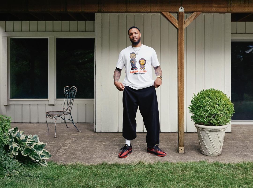 Adult male poses outdoors on a concrete patio, wearing a t-shirt, baggy pants, and athletic shoes, next to a potted green bush.