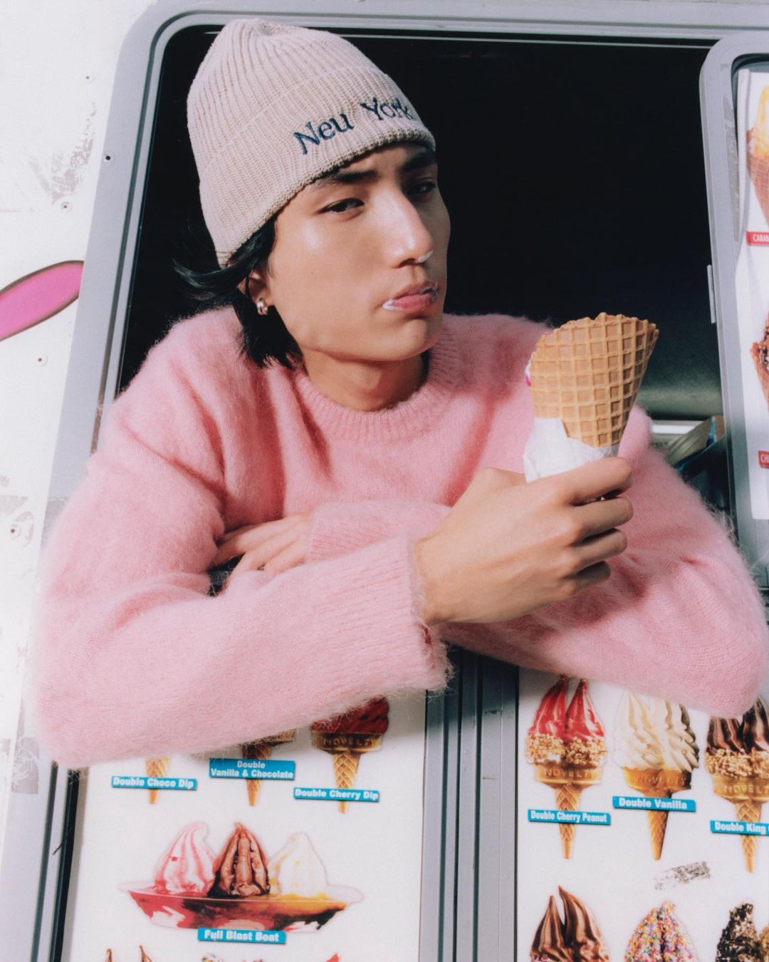 Young adult Asian man in a pink sweater and beanie holds a waffle cone while leaning out of an ice cream truck window.