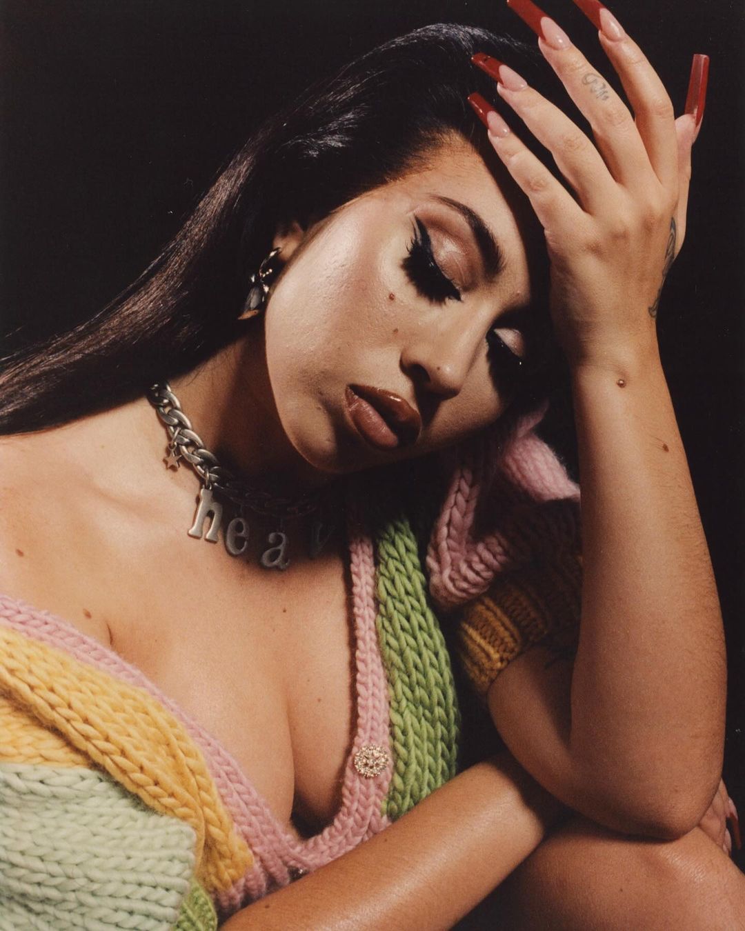 Close-up portrait of a woman with long dark hair, long red nails, and a colourful knit cardigan against a dark background.