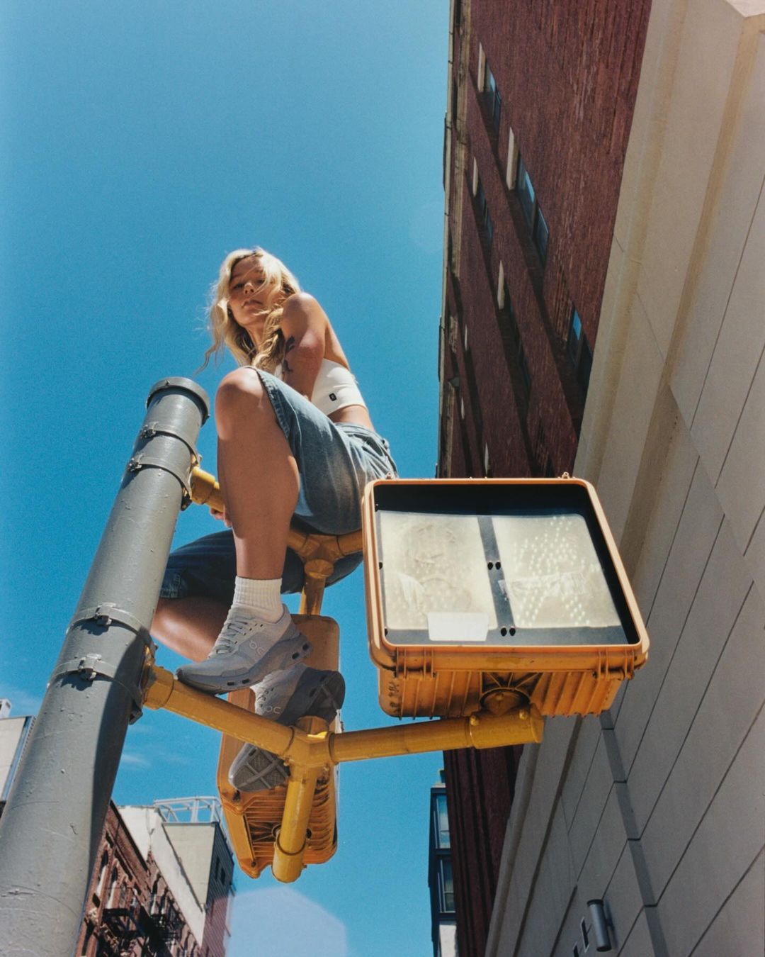 Woman sitting on traffic light in city. Low angle captures youthful energy. Buildings and blue sky form a striking backdrop.