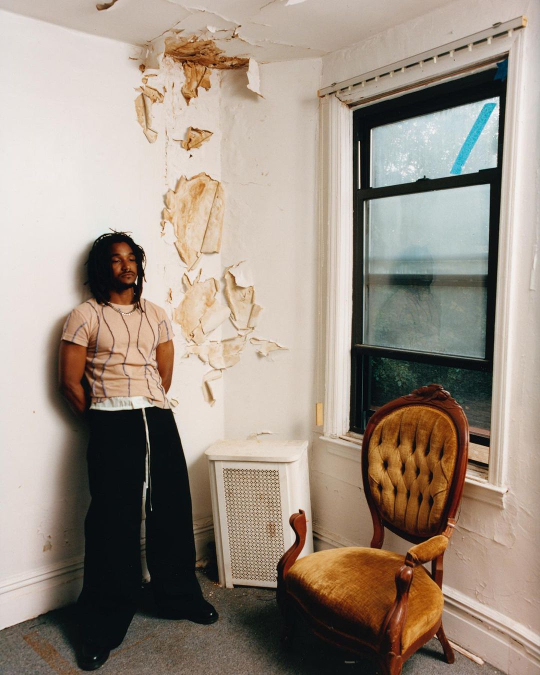 Adult male posing in a room with damaged walls, wearing a beige shirt and black pants, with a chair and radiator in the background.
