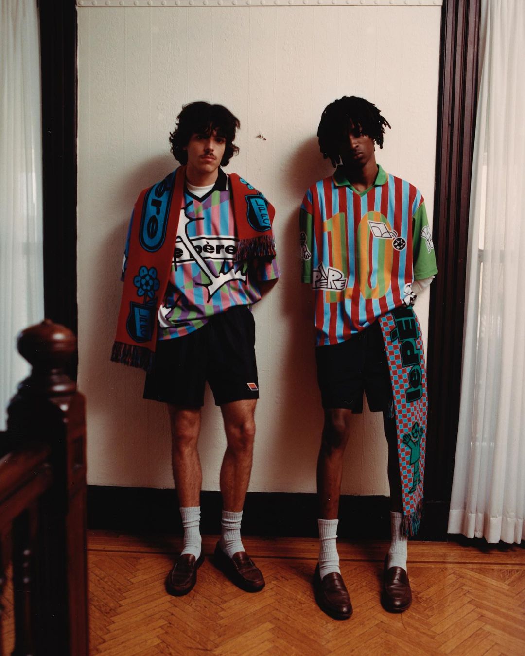 Two adult male fashion models in patterned shirts and shorts with casual loafers standing in an indoor setting near a dark wood banister.