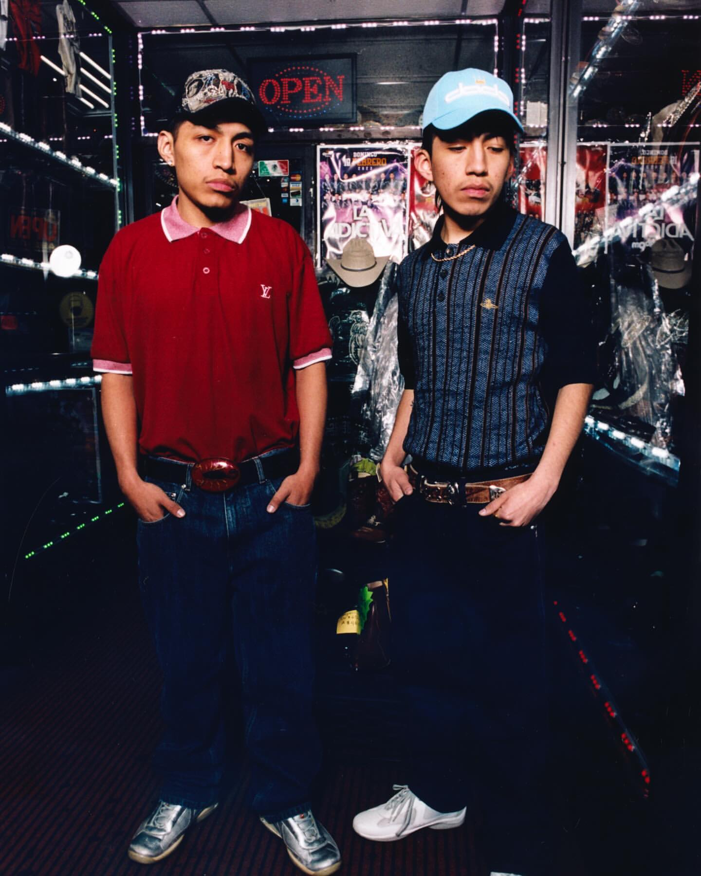 Two young men in casual attire pose indoors in a retail environment with cowboy hats and neon 'OPEN' sign in the background.