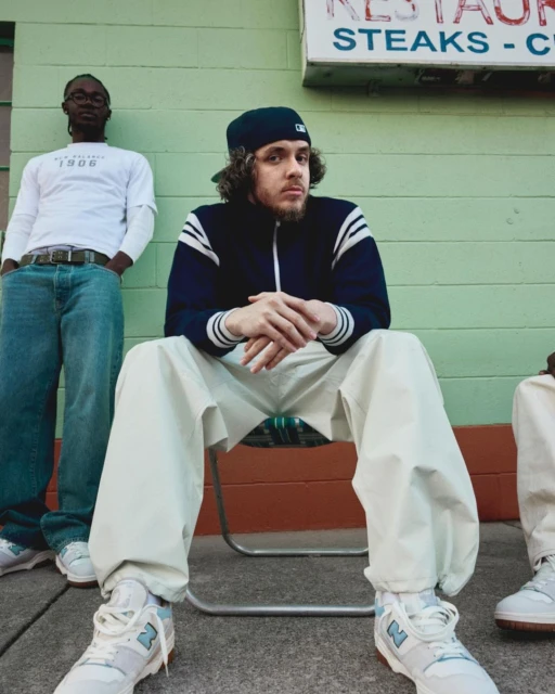 Three people in streetwear pose in front of a building with light green siding and a restaurant sign. One person sits in a chair.