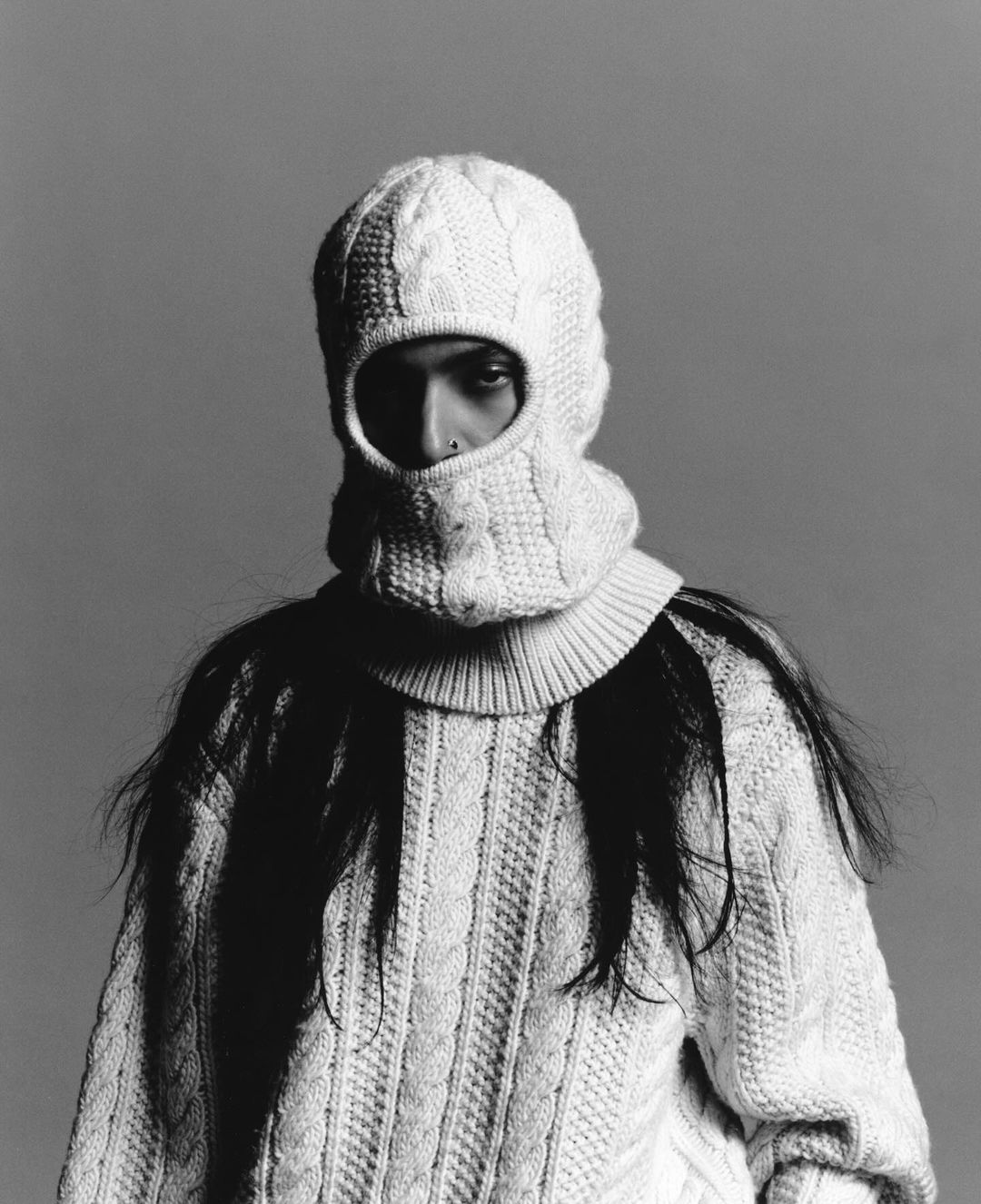 A person wears a white knitted balaclava and matching sweater in a studio portrait against a grey backdrop. The portrait is in black and white.