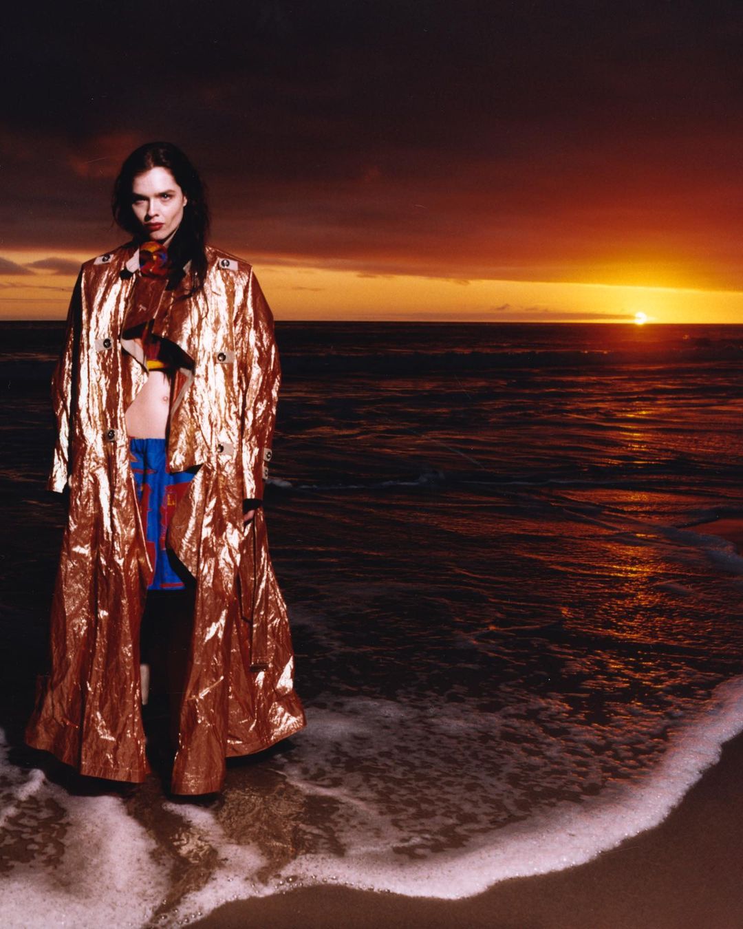 Brunette fashion model wearing a gold coat stands on a sandy beach with water lapping at her feet, with a vibrant sunset background.