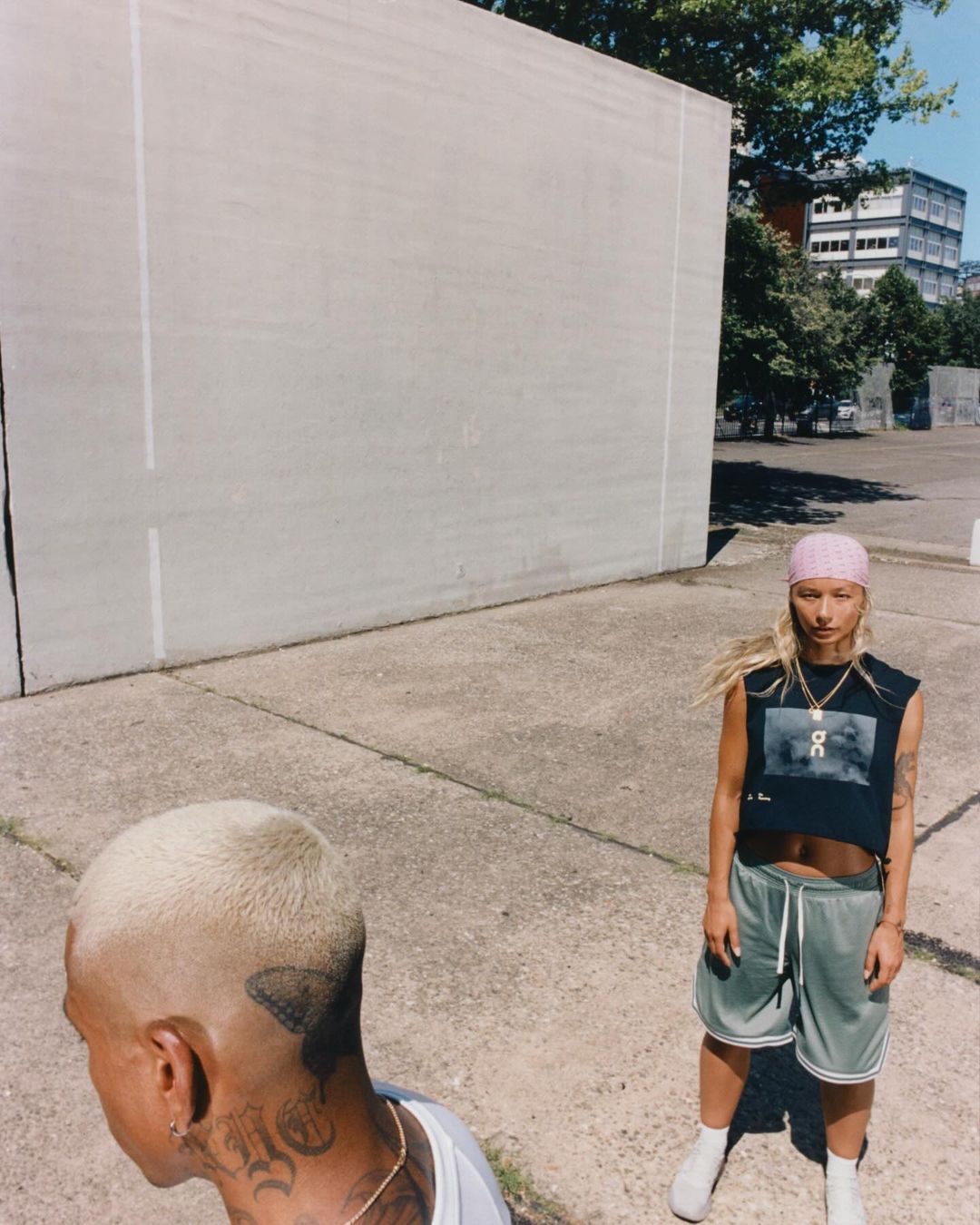 Two people in an outdoor setting with neutral tones. One person faces forward, wearing a crop top and athletic shorts; the other is partially visible.