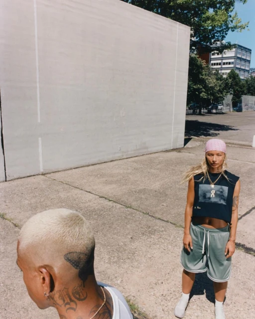 Two people in an outdoor setting with neutral tones. One person faces forward, wearing a crop top and athletic shorts; the other is partially visible.