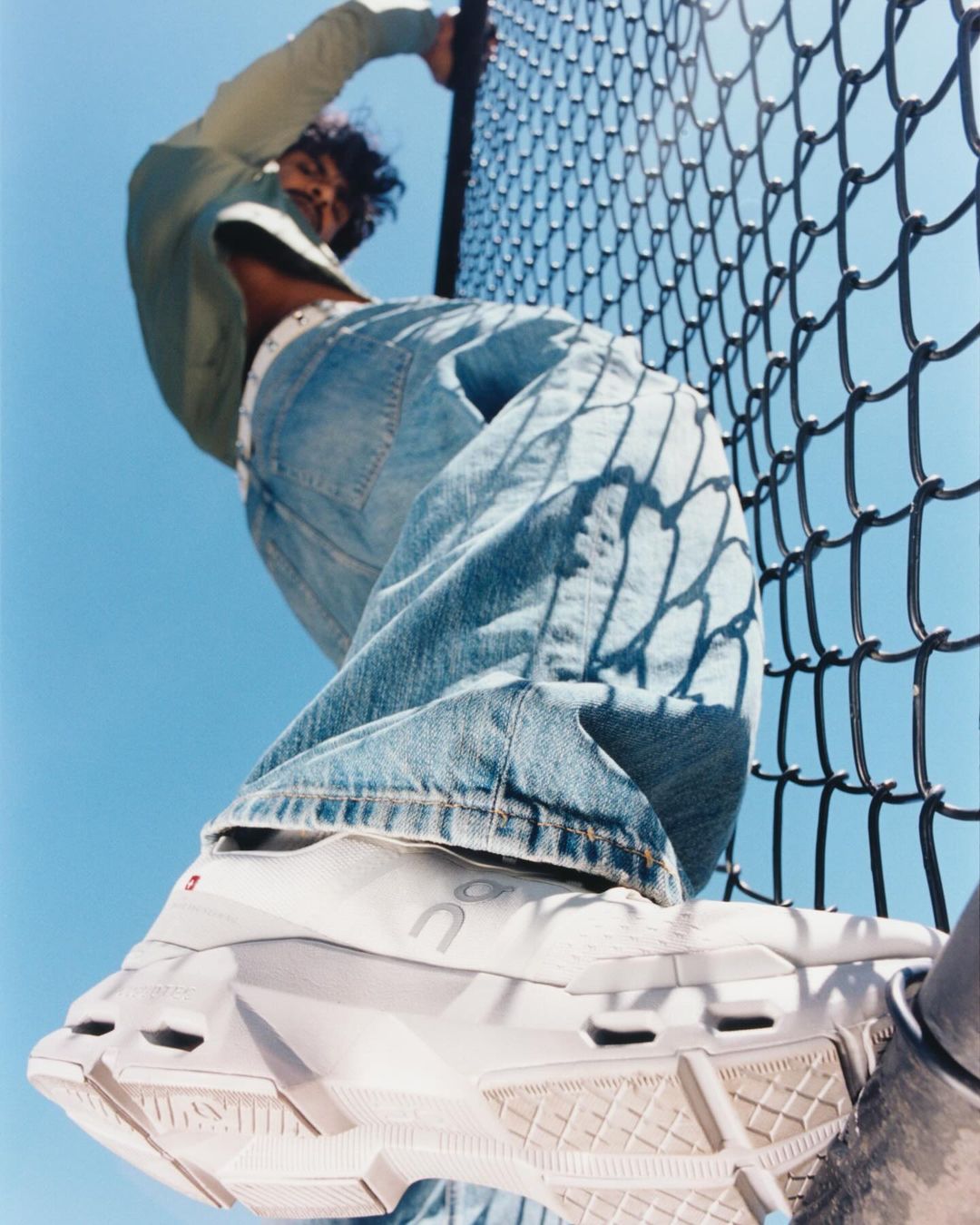 Eye-level view of a person leaning on a chain-link fence in casual clothes and white athletic shoes against a clear blue sky.
