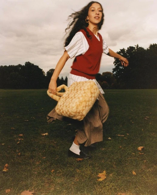 A young adult woman in a preppy outfit, including a vest and oversized bag, strides across a green lawn with scattered fallen leaves.