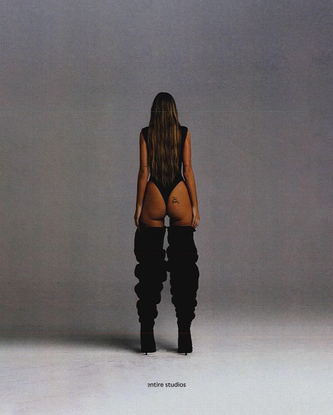 A woman is facing away from the camera wearing a black bodysuit and thigh-high boots in a studio setting with grey background.