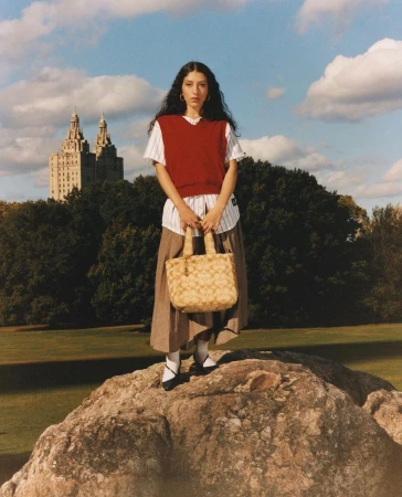 Woman standing on a rock holding a beige handbag, wearing a red sweater vest, striped shirt, and pleated skirt in a park.