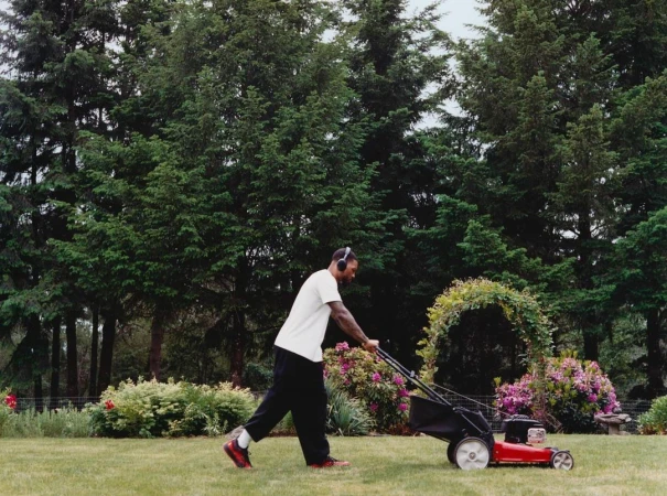 An adult man mowing a lawn in a yard with green grass, a red lawnmower, headphones, wearing a white shirt and black pants. Tall green trees.
