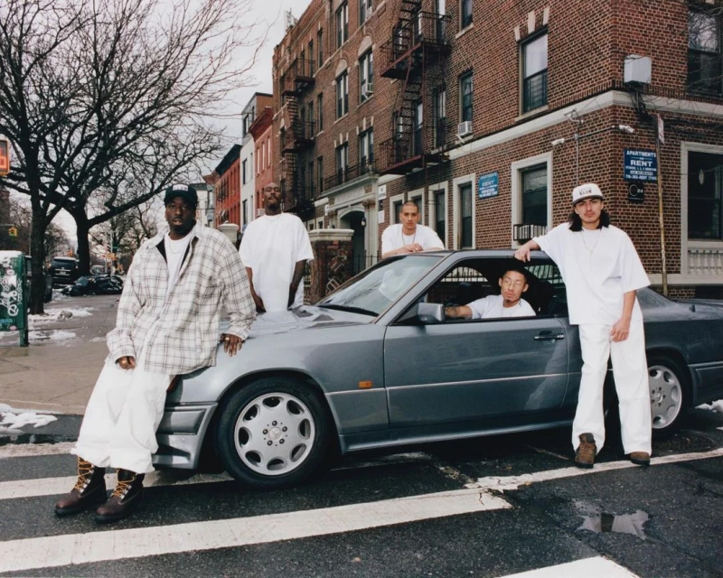 Group of five people and a grey car on a city street with brick buildings and bare trees during the day, wearing white clothing and hats.