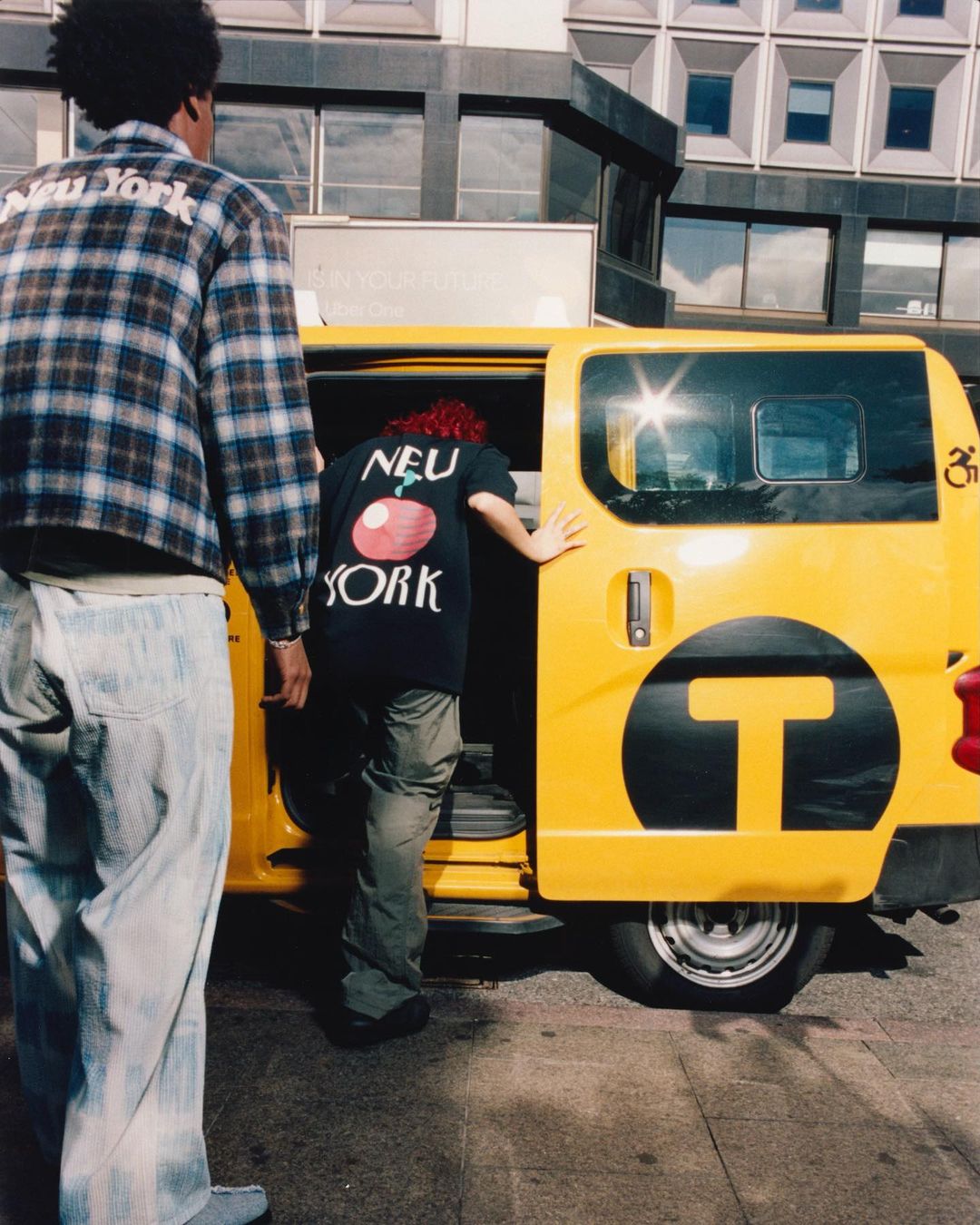 Two people stand near a yellow taxi with 'New York' graphics. One person wears a plaid jacket, the other a printed shirt.