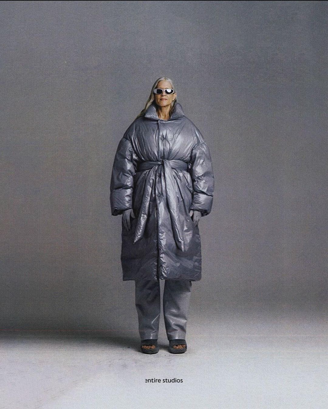 Mature adult woman in an oversized puffer coat, sunglasses, and gray matching set poses in a photo studio setting. Fashion editorial.