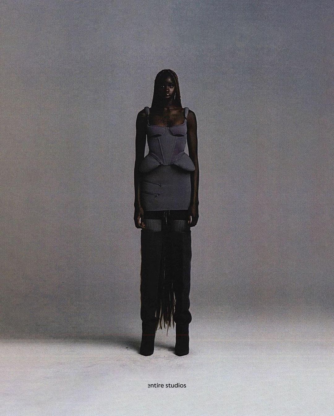 A striking full shot captures a Black woman in a grey corset-style dress, set against a smooth grey backdrop in a studio setting.
